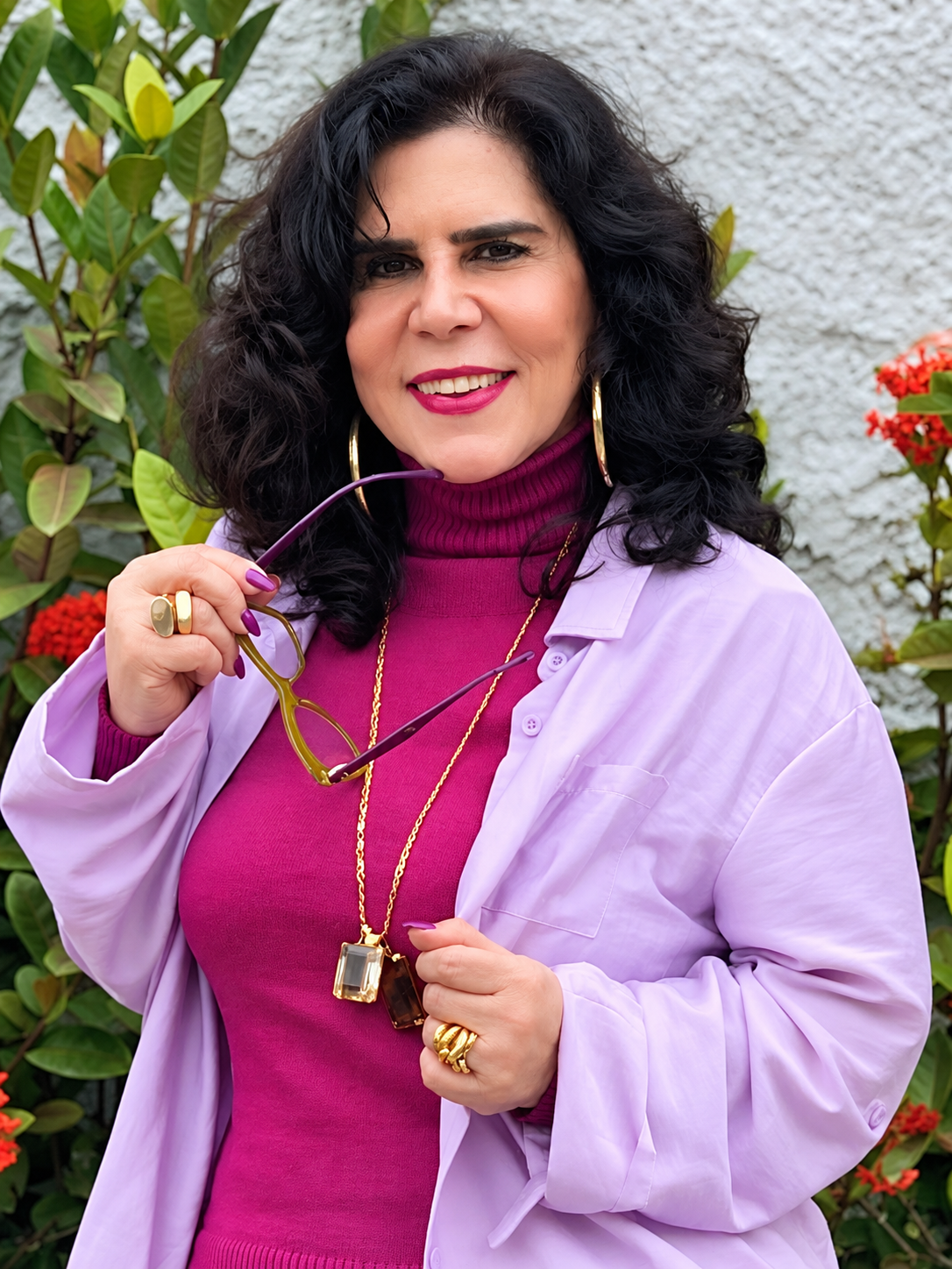 Retrato de Fátima Castro em ambiente externo, com flores ao fundo, usando roupa lilás e segurando óculos.